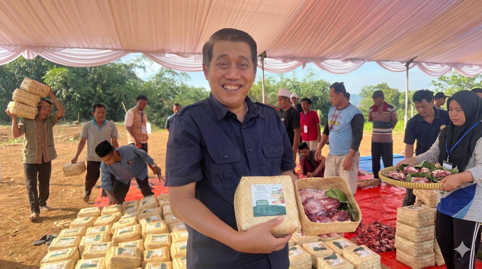 Panitia memegang besek berisi daging kurban di lokasi pembagian, tumpukan besek dan warga di sekitarnya