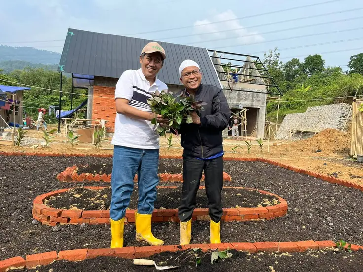 Dua orang memegang bibit ubi ungu di bedeng kebun kawasan, bangunan yang sedang dibangun tampak di belakang