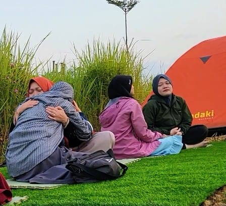 Peserta kegiatan duduk di rumput dekat tenda, dua di antaranya berpelukan